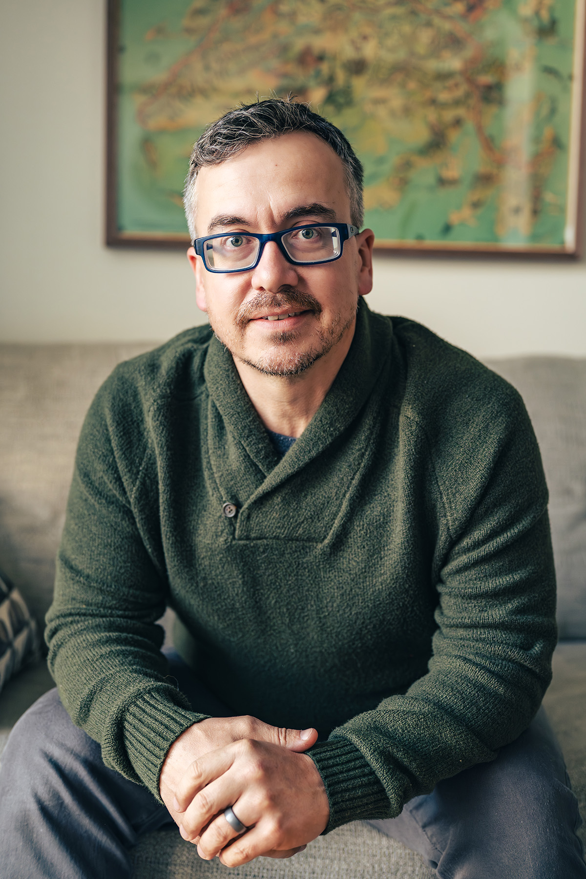 Photo of Desmond - a man wearing blue plastic glasses, a green sweater, with short hair and stubby facial hair smiling 
