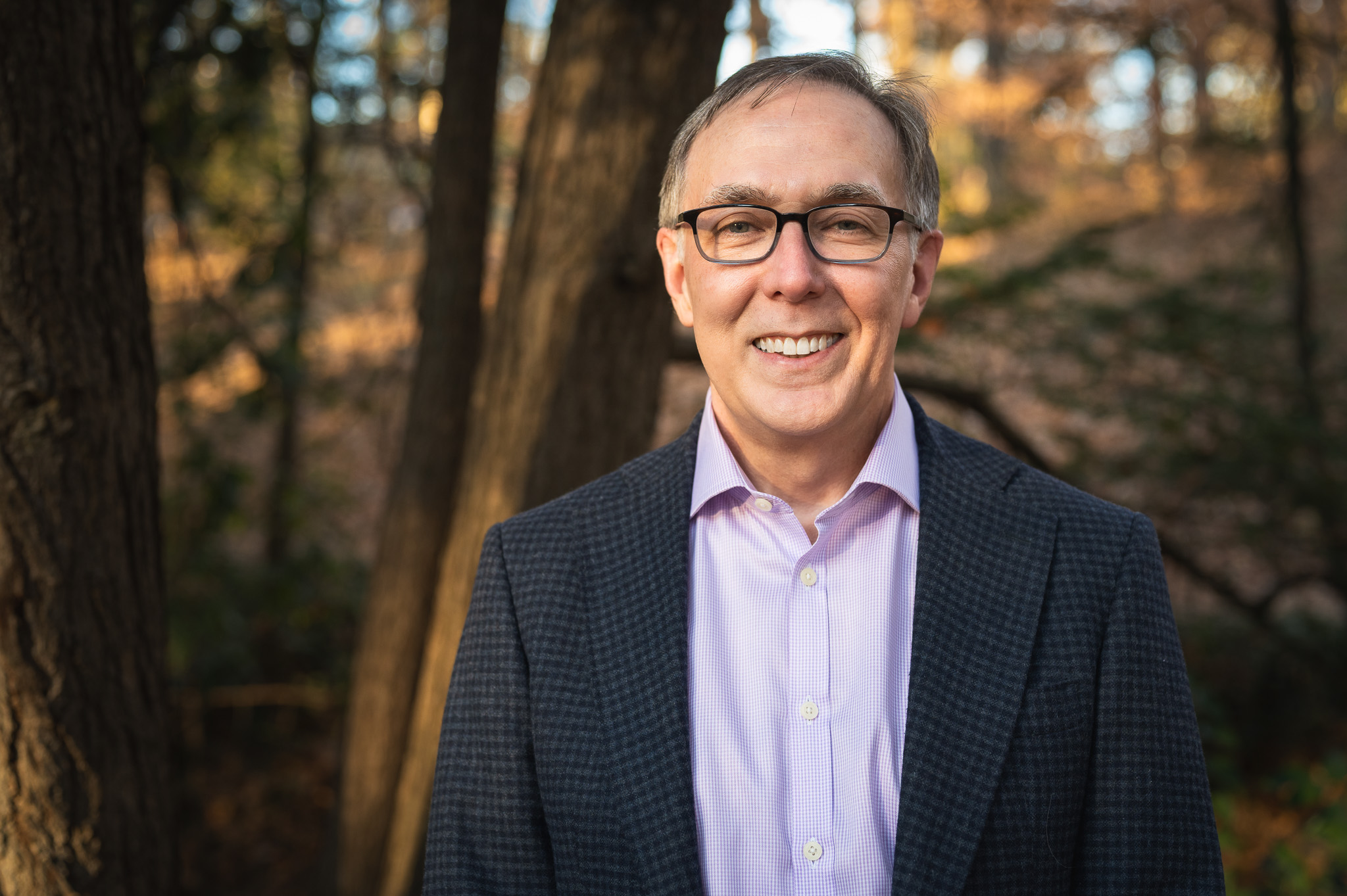 A headshot/profile photo of a gentleman who is smiling and wearing a blue blazer and violet shirt.